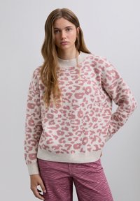 Junge Frau mit langen Haaren, die einen rosa und weißen Leopardenmuster-Pullover und gemusterte rosa Hosen trägt, steht vor einem einfarbigen Hintergrund.