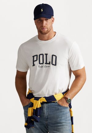 Witte katoenen t-shirt met "POLO" grafiek, gecombineerd met een marineblauwe pet en een gele/marineblauwe gestreepte sweater om de taille. Draag het met blauwe jeans.