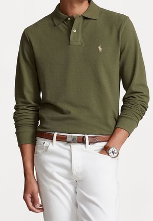 Man draagt een olijfgroen poloshirt met lange mouwen en een klein logo op de borst, witte broek, bruine lederen riem en een horloge met een bruine band.