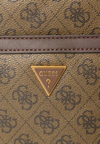 Brązowa torebka syntetyczna z wzorem i teksturowanym wykończeniem, z pozłacaną trójkątną plakietką z napisem „GUESS” oraz ciemnym skórzanym akcentem.