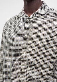 Homme portant une chemise à carreaux beige et noire à boutons, avec une poche avant et un col pointu, montré du cou au milieu du torse.