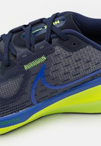 Primo piano di una scarpa da corsa blu navy e grigia con logo swoosh blu, lacci blu navy e dettaglio della suola giallo-verde brillante.