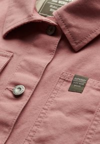 Camicia di stoffa rosa con design a bottoni, caratterizzata da un bottone in metallo e un'etichetta a patch che recita "Superdry Work Wear". Materiale testurizzato.
