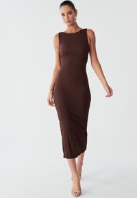 Brun ribstrikket midikjole med bådformet halsudskæring, tætsiddende silhuet og detaljer med rynkninger i siden. Modellen bærer beige stropper på hælene.