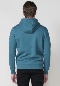 Felpa con cappuccio di colore blu teal, realizzata in tessuto morbido. Presenta una tasca a marsupio, polsini e orlo a coste. Indossata con pantaloni neri.