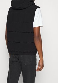 Schwarze Pufferweste mit Kapuze, ausgestattet mit horizontalen Steppnähten. Über einem weißen T-Shirt getragen und kombiniert mit grauen Jeans.
