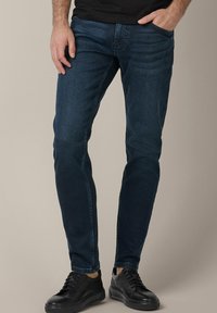 Mrocznie niebieskie dżinsy o slim fit, z delikatnym efektem sprania, klasycznym pięciokomorowym designem, w połączeniu z czarnymi trampkami.