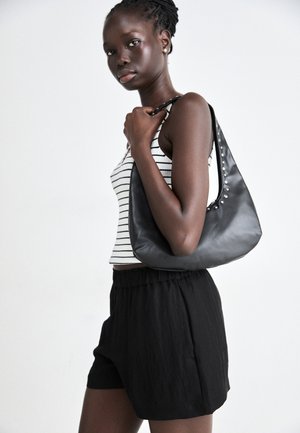 Schwarze Lederhandtasche mit gebogener Form, versehen mit silbernen Nieten entlang der Naht. Das Modell trägt ein gestreiftes Tanktop und schwarze Shorts.