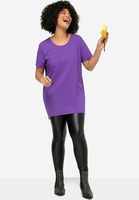 Paarse tuniek met korte mouwen gecombineerd met zwarte synthetische leggings en gestikte zwarte enkelboots. Het shirt heeft een relaxte pasvorm en een ronde halslijn.
