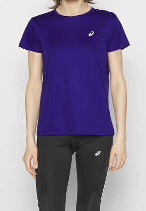 Personne portant un t-shirt de sport violet à manches courtes et des leggings noirs, tous deux avec des logos blancs ASICS, debout devant un fond uni.