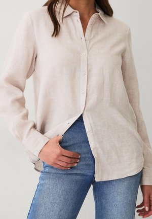 Femme portant une chemise en lin beige clair boutonnée avec un jean bleu, une main dans la poche du jean, visible du cou aux hanches.