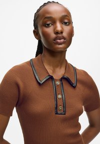 Pruun soonik polo särk musta äärega kaelusega, mille esiosas on kolm kuldset nuppu. Õhuke ja kehaümberlükatav disain ning tekstuuriga kangas.