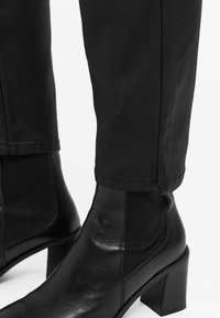 Bottes noires en cuir au niveau de la cheville avec un talon large, dotées de panneaux élastiques sur les côtés et d'une texture lisse, associées à un pantalon noir fuselé.