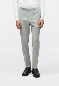 Calças tailor cinzas claras com um corte slim, apresentando uma textura suave e bolsos laterais, combinadas com sapatos pretos de slip-on.