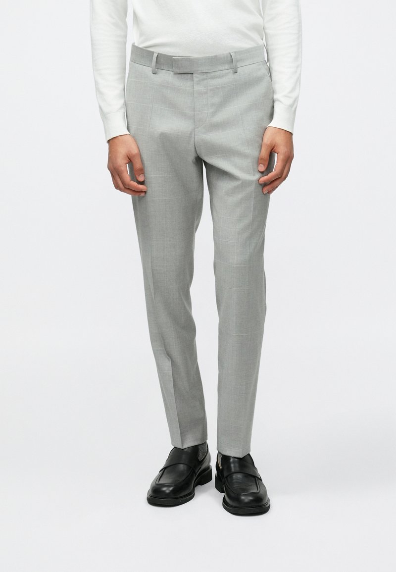 Calças tailor cinzas claras com um corte slim, apresentando uma textura suave e bolsos laterais, combinadas com sapatos pretos de slip-on.