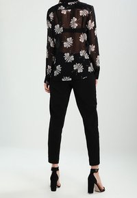 Blusa nera trasparente con stampa floreale, abbinata a pantaloni neri su misura. La modella indossa tacchi neri con cinturini che mettono in risalto un design elegante.