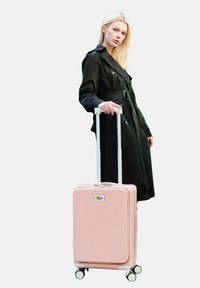 Rosa Hartschalenkoffer mit vertikaler Rippentextur, weißen Reißverschlüssen und Rädern, begleitet von einem Modell in einem schwarzen Trenchcoat.