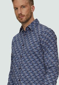 Niebieska koszula w wzory z geometrycznym designem, z kołnierzykiem typu button-down i brązowymi guzikami, wykonana z lekkiego materiału o gładkiej fakturze.
