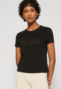 Camiseta negra de manga corta con un logo texturizado de "GUESS LOS ÁNGELES" en un acabado brillante. Material de algodón suave, diseño de cuello redondo.