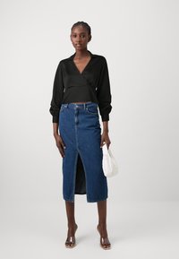 Top nero avvolgente con maniche lunghe, abbinato a una gonna di jeans aderente con uno spacco frontale. Accessorio: piccola borsa bianca.