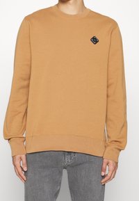 Hnědá mikina s kulatým výstřihem, dlouhými rukávy a černou nášivkou ve tvaru diamantu na hrudi. Měkký materiál a žebrované manžety.