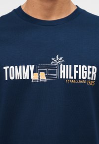 Μπλε ναυτικό πουκάμισο με λευκό και πορτοκαλί κείμενο "Tommy Hilfiger Established 1985", δύο κίτρινες ξαπλώστρες, ένας φοίνικας και μια εικόνα καμπίνας.