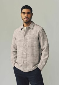 Textured beige jacket with a button-up front, collar, and side pockets; worn over a light-colored shirt. Dark pants complete the look.