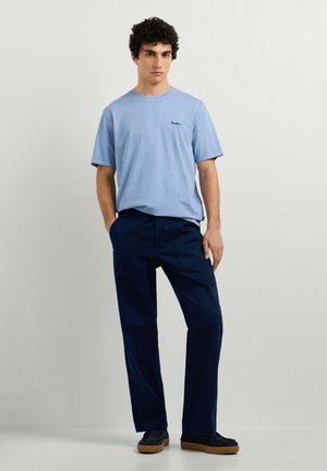 Jonge man staand met handen in de zakken, draagt een lichtblauw T-shirt en donkerblauwe broek tegen een effen achtergrond.