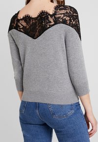 Pull gris avec une encolure en dentelle noire, manches trois-quarts et ourlet côtelé. La dentelle présente un motif floral et des bords festonnés.