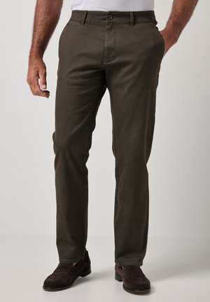 Hombre con pantalones de pierna recta en color oliva oscuro con bolsillos delanteros y traseros y mocasines de ante marrones, de pie con una mano en el bolsillo.