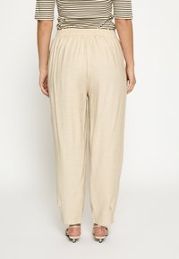 Beežid elastse vööosaga püksid, mille lõige on mugav ja lõtv, omades siledat tekstuuri ja peent läiget, kantud avatud varbaga kontsakingadega.