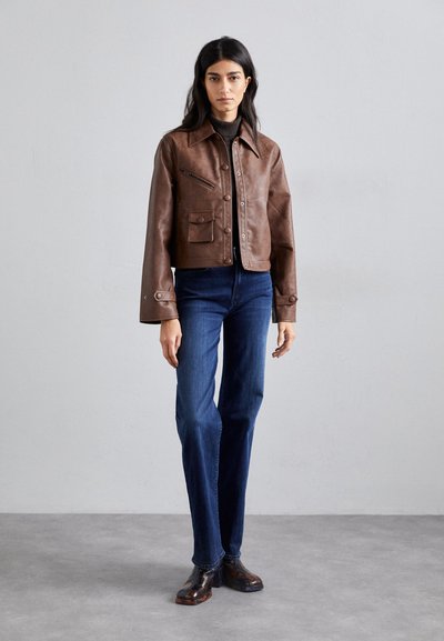 Chaqueta corta de cuero marrón con bolsillos frontales y cierre de botón, combinada con jeans rectos azules y botas oscuras con patrón.