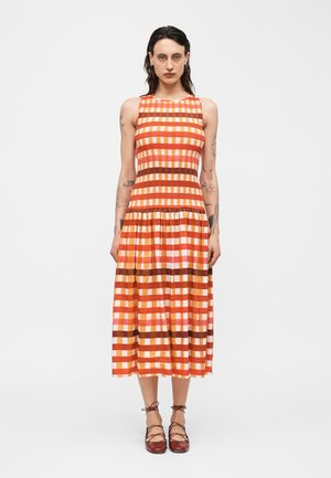 Femme tatouée portant une robe midi sans manches à carreaux orange, rose et marron, ainsi que des ballerines marron à lacets, debout devant un fond blanc.