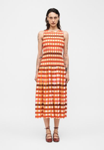 Femme tatouée portant une robe midi sans manches à carreaux orange, rose et marron, ainsi que des ballerines marron à lacets, debout devant un fond blanc.