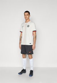 Maglia sportiva bianca a maniche corte, con logo, dettagli rossi e verdi, abbinata a pantaloni corti neri, calze bianche e scarpette nere.
