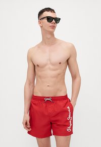 Červené plavky s pružným pásom na šnúrku, s bielym logom "Pepe Jeans" na ľavej strane. Hladká látka, dĺžka po kolená a bez vzorov.