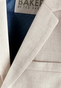 Blazer beige in tessuto testurizzato; presenta rever a punta, una tasca sul petto e una fodera blu a fantasia con etichetta del marchio.