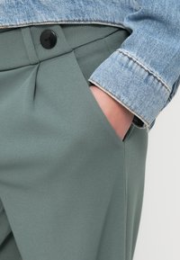 Hellgrüne, maßgeschneiderte Hose mit Falten und Seitentaschen, kombiniert mit einer Jeansjacke aus blau strukturiertem Stoff und Knopfdetail.