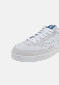 Zapatilla blanca y gris con un upper de cuero suave y acentos de ante. Presenta perforaciones en el lateral y cordones planos blancos. Logo azul en la lengüeta.