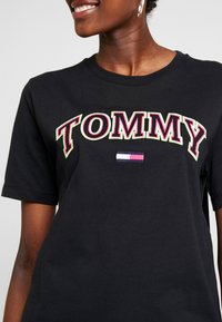 Camiseta de algodón negra con el texto bordado "TOMMY" en rosa y verde, con un pequeño detalle de bandera tricolor horizontal debajo. Manga corta.