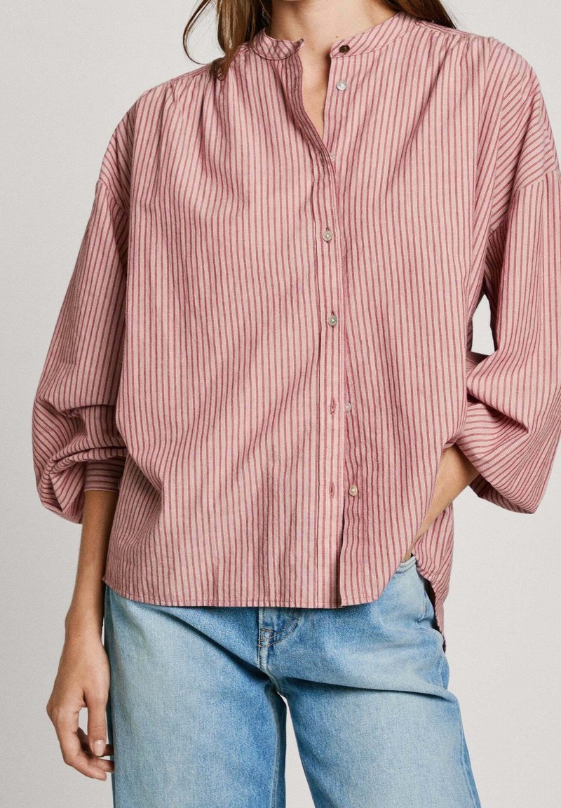 Femme portant un chemisier ample à rayures verticales roses et rouges, boutonné, avec de longues manches bouffantes et un jean bleu clair. Une main dans la poche.