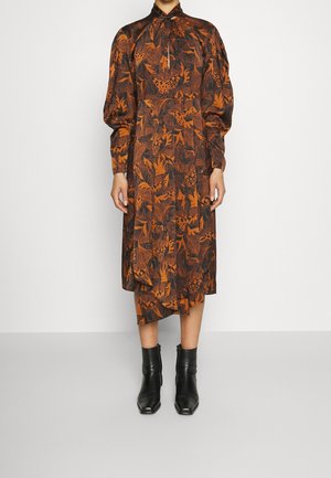Kobieta w midi sukience w rdzawo-pomarańczowy i czarny wzór liści, z bufkami na rękawach, wysokim kołnierzem i czarnymi botkami za kostkę.