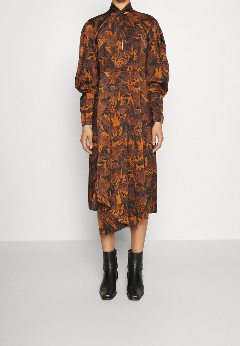 Femme portant une robe midi à motifs feuilles orange rouille et noire, manches bouffantes, col montant et bottines noires.