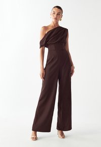 Rjava kombinacija z eno naramnico, s prilagojenim korzetom, tekočo draperijo in širokimi hlačnicami. Gladek material s sijočo teksturo, združen z sandali s trakovi.