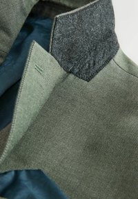 Nahaufnahme eines Jackenkragens mit grünem Strukturstoff, einem grauen Filz-Unterkragen und einem blau geblümten Futter innen.