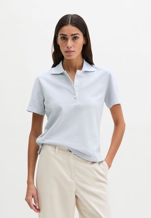 Vrouw die een lichtblauw korte mouwen poloshirt draagt, ingestopt in beige broek met hoge taille, staand met één hand in de zak.