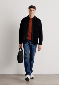 Giacca camicia in velluto corduroy nera sopra un maglione arancione a coste, jeans blu e sneakers bianche. Porta una borsa nera con manico.