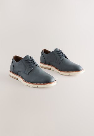 Scarpe eleganti da uomo in pelle blu con finitura liscia, caratterizzate da punta arrotondata, chiusura frontale con lacci e suola bianca con dettagli marroni a contrasto.