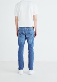 Blå denimjeans med rak passform, fem fickor och en tan läderlapp vid midjan, tillsammans med en vit t-shirt med rund halsringning.