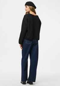 Maglione nero cropped con maniche ampie, abbinato a jeans blu scuro a vita alta. Il modello indossa un berretto nero, mostrando uno stile casual e rilassato.
